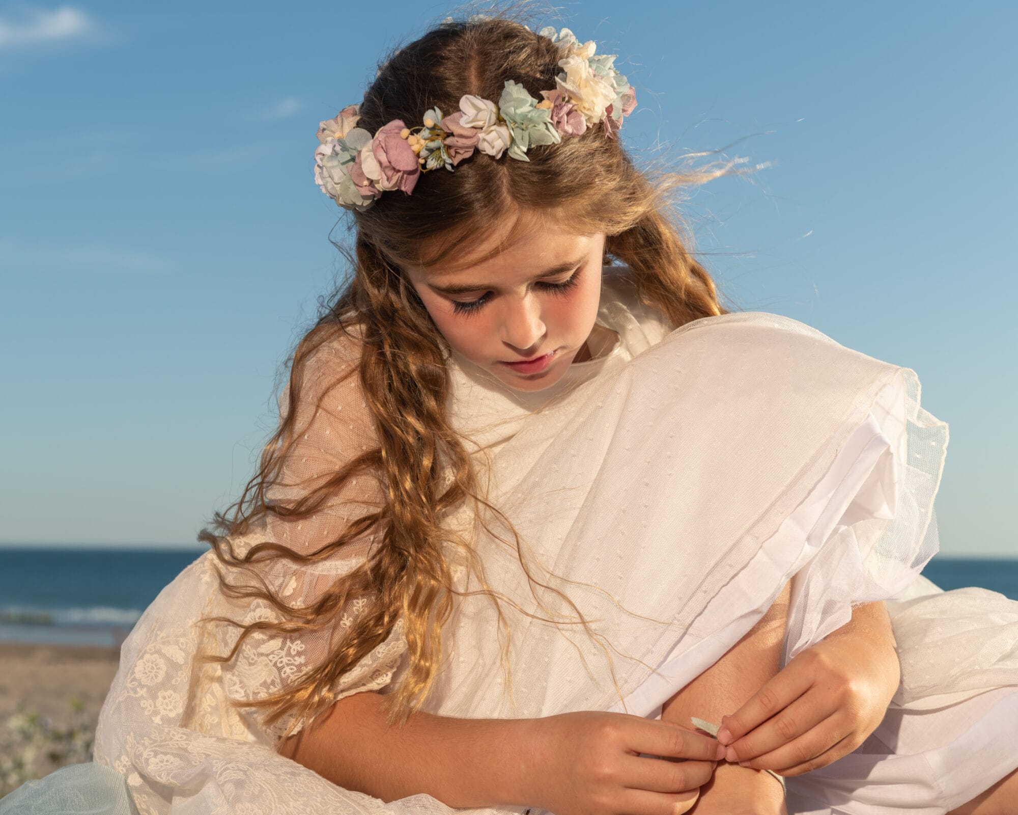Niña con corona de flores y vestido de comunión blanco en la playa bajo la luz del sol en Andalucía
