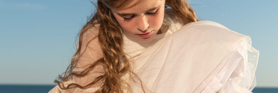 Niña con corona de flores y vestido de comunión blanco en la playa bajo la luz del sol en Andalucía