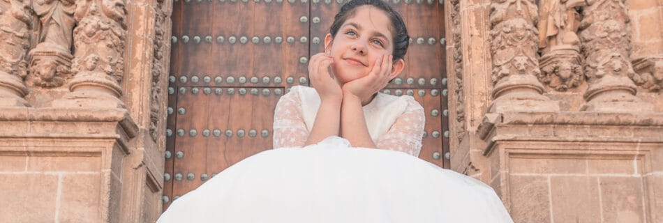 Niña con vestido de comunión blanco posando frente a la majestuosa puerta tallada de una iglesia histórica.