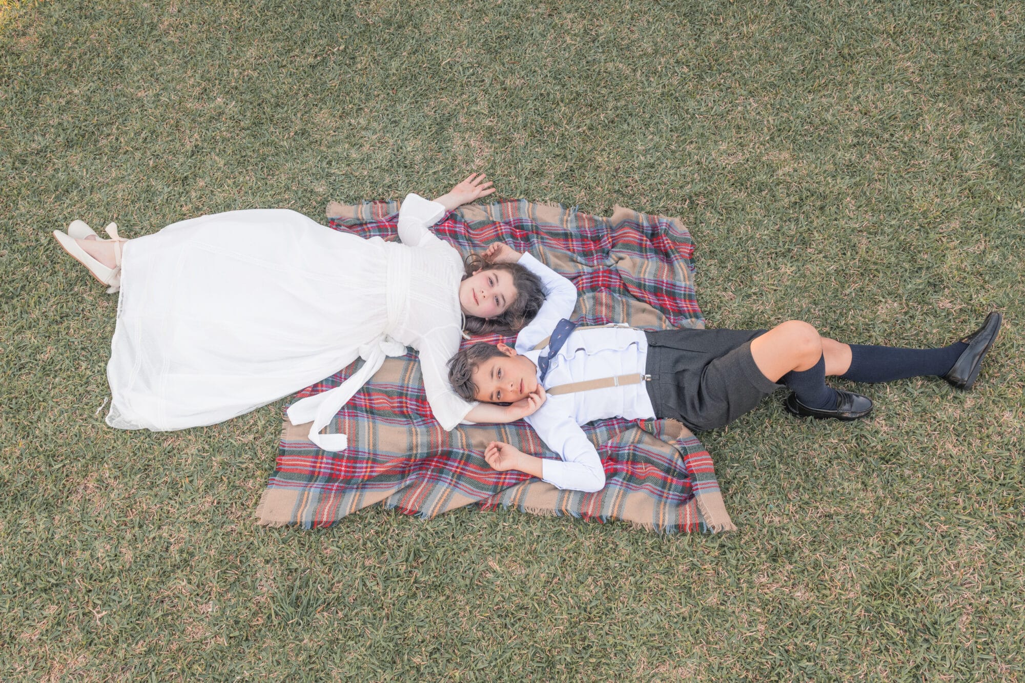 Niña con vestido de comunión y niño con tirantes acostados en una manta en el césped en Jerez