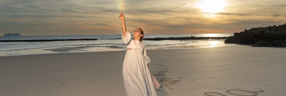Niña vestida de comunión en la playa al atardecer, levantando un brazo hacia el cielo queriendo tocar el arco iris mientras su nombre, “Carla”, está escrito en la arena.