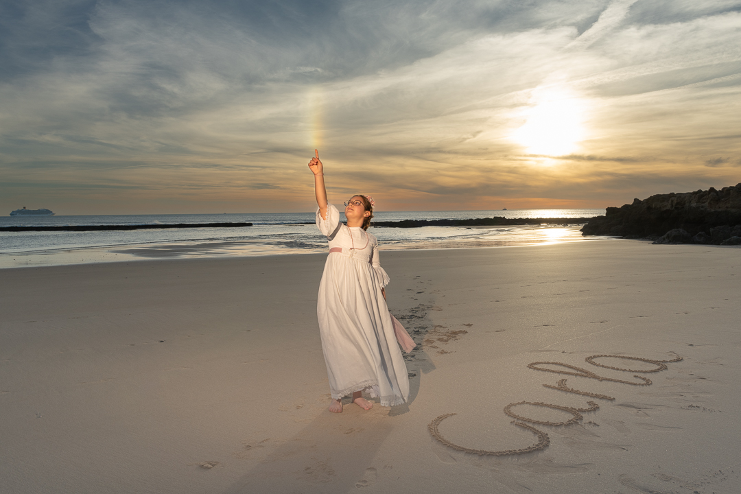 Niña vestida de comunión en la playa al atardecer, levantando un brazo hacia el cielo queriendo tocar el arco iris mientras su nombre, “Carla”, está escrito en la arena.