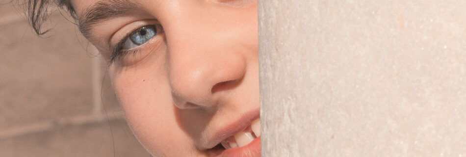 Retrato de un niño asomándose detrás de una pared, mostrando una sonrisa y destacando sus ojos claros en un entorno de piedra.